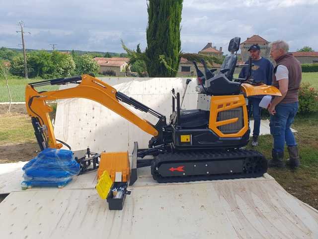 A yellow mini excavator and attachments on a wooden base with two people nearby.